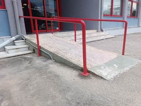 Τσιμεντένια ράμπα με κόκκινο κιγκλίδωμα, οδηγεί σε τέσσερα σκαλοπάτια και γυάλινη αυτόματη θύρα.