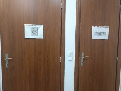 Δύο καφέ πόρτες σε εσωτερικό χώρο, η αριστερή φέρει το αναπηρικό σήμα, η δεξιά φέρει την ένδειξη 'toilets' στα αγγλικά.
