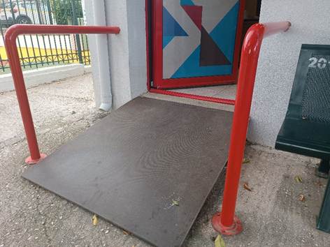 Μικρή ράμπα σε γκρι κτίριο με κόκκινο κιγκλίδωμα και κόκκινη πόρτα.