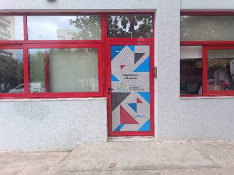 Γκρι κτίριο με κόκκινες λεπτομέρειες, είσοδος που αναγράφει Δημοτολόγιο Ληξιαρχείο.